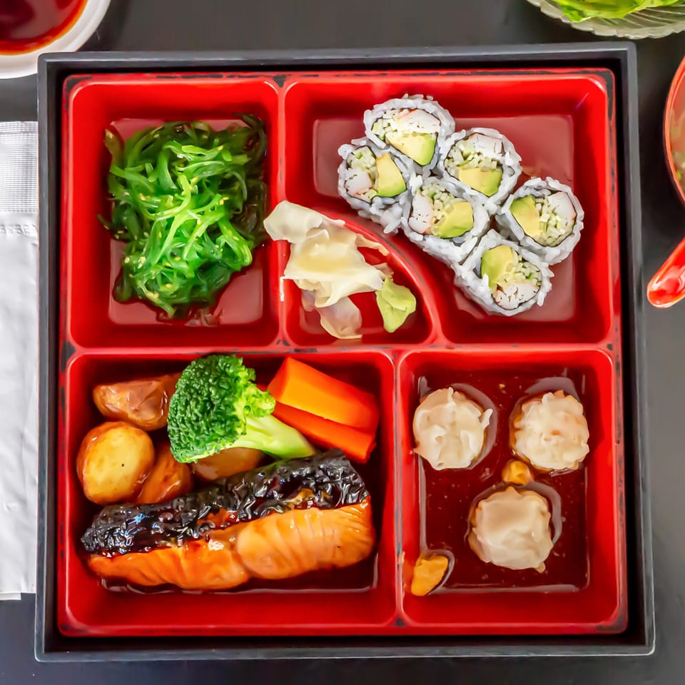 A Koiso Japanese Restaurant bento box with salmon, sushi, seaweed salad, and dumplings.