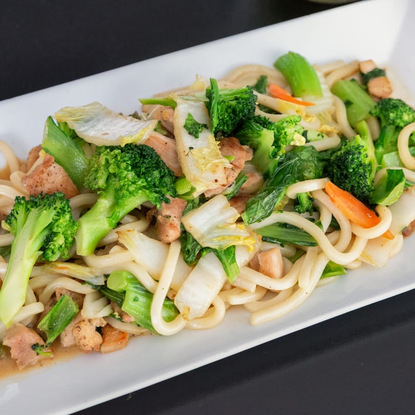 Japanese udon noodles with chicken, broccoli, bok choy, and carrots on a white rectangular plate.