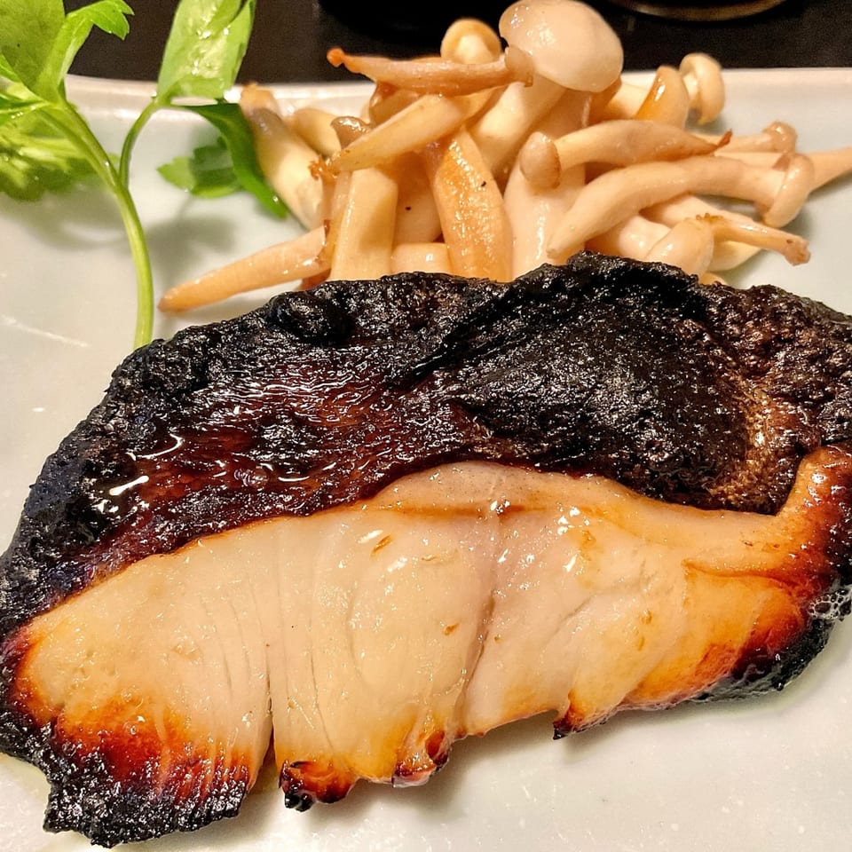 Grilled black cod with a side of sautéed mushrooms and a green garnish on a white plate.
