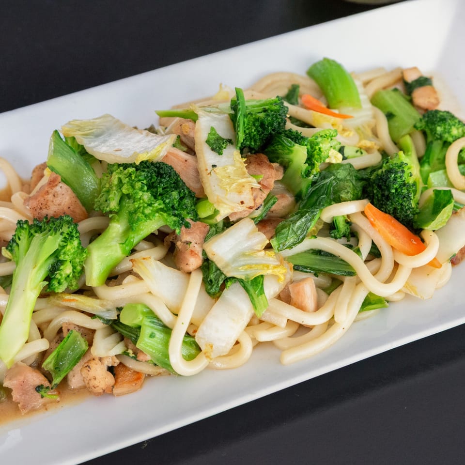 Japanese udon noodles with chicken, broccoli, bok choy, and carrots on a white rectangular plate.